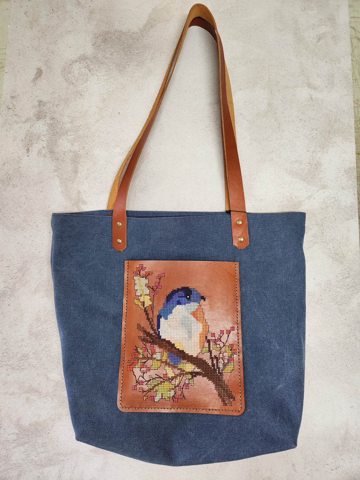 Cross-stitch tote bag with a bird design, blue fabric and tan leather handles